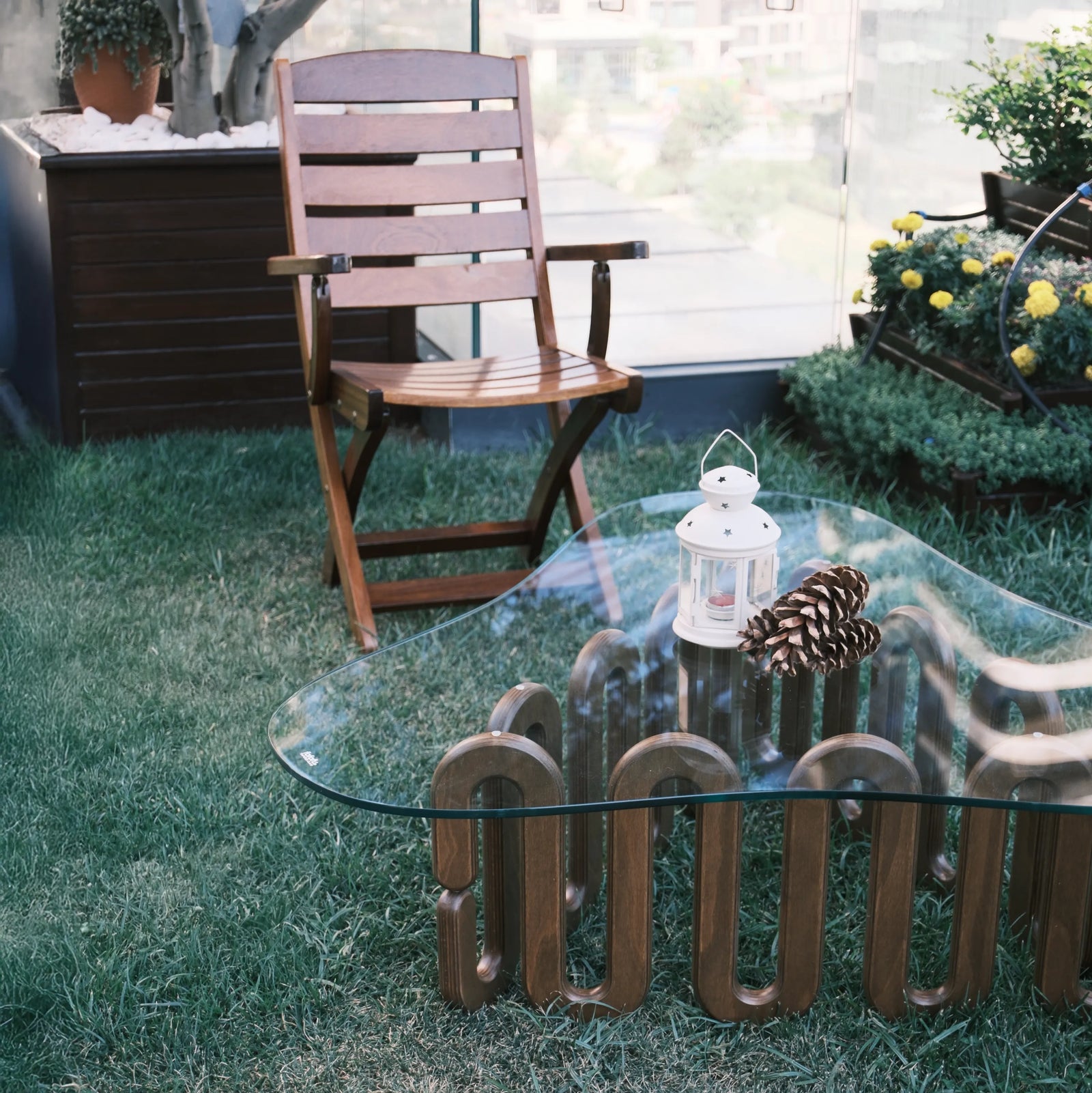 Moderner Couchtisch mit Glasplatte und geschwungenem Holzfuß im Garten neben dunklem Rattanstuhl vor weißer Hauswand
