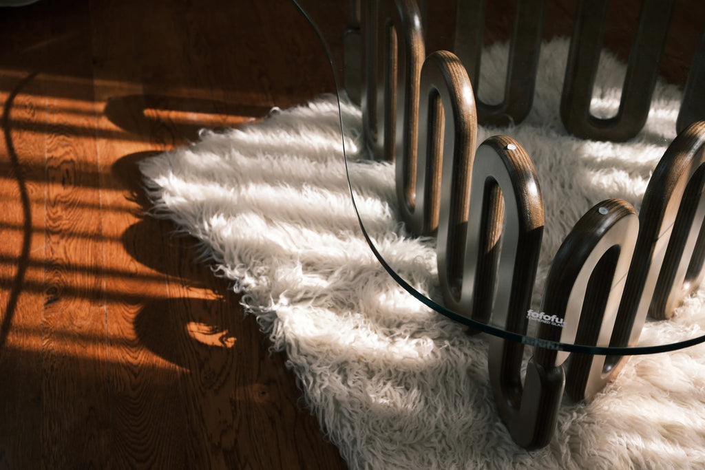 Detailaufnahme eines kunstvoll geschwungenen Holzfußes mit Glasplatte auf weißem Teppich, modernes Design im Sonnenlicht auf Parkett.