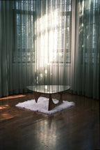 Design-Couchtisch mit Glasplatte und Holzgestell auf weißem Teppich in hellem Raum mit Parkettboden und lichtdurchfluteten Vorhängen.