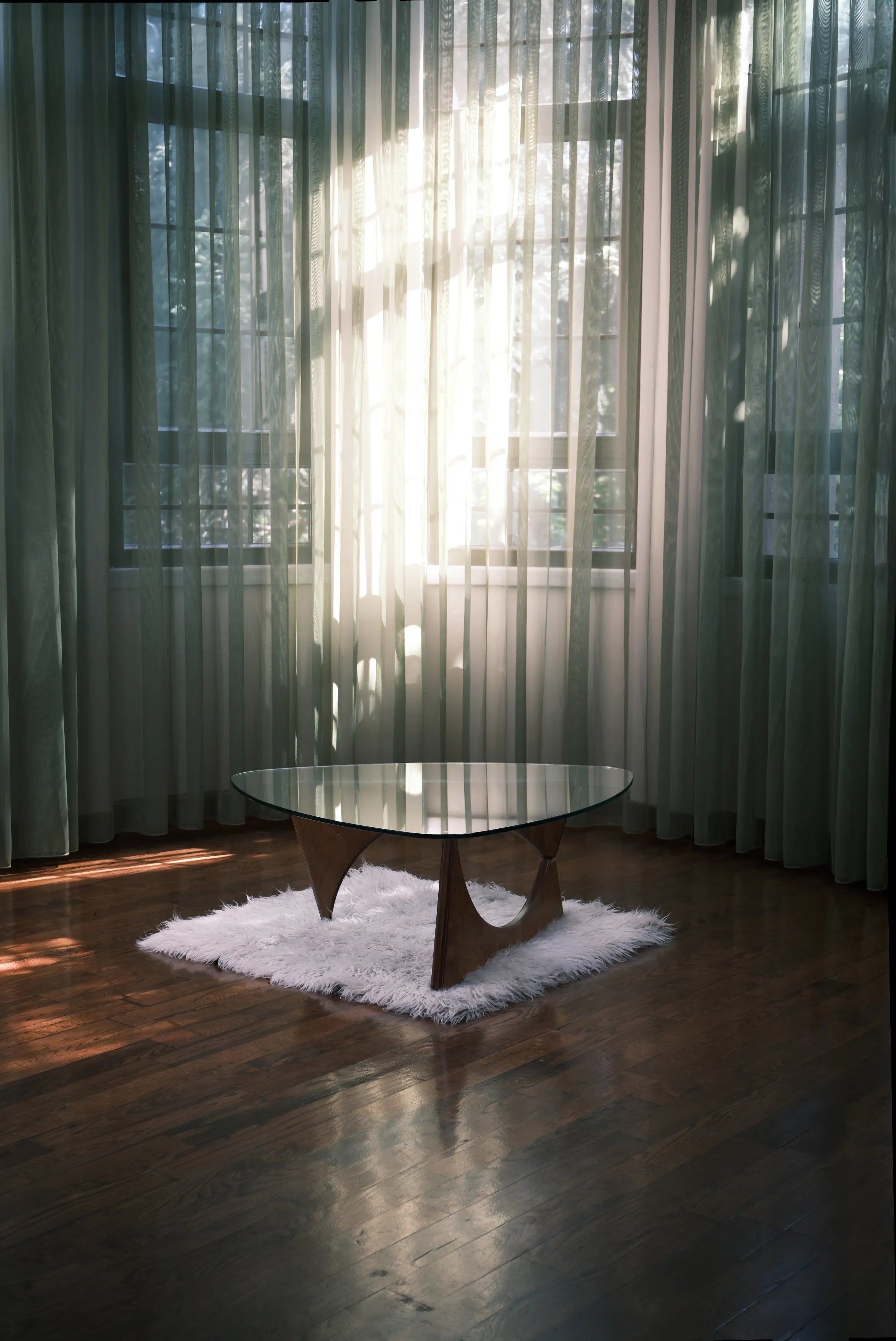 Design-Couchtisch mit Glasplatte und Holzgestell auf weißem Teppich in hellem Raum mit Parkettboden und lichtdurchfluteten Vorhängen.