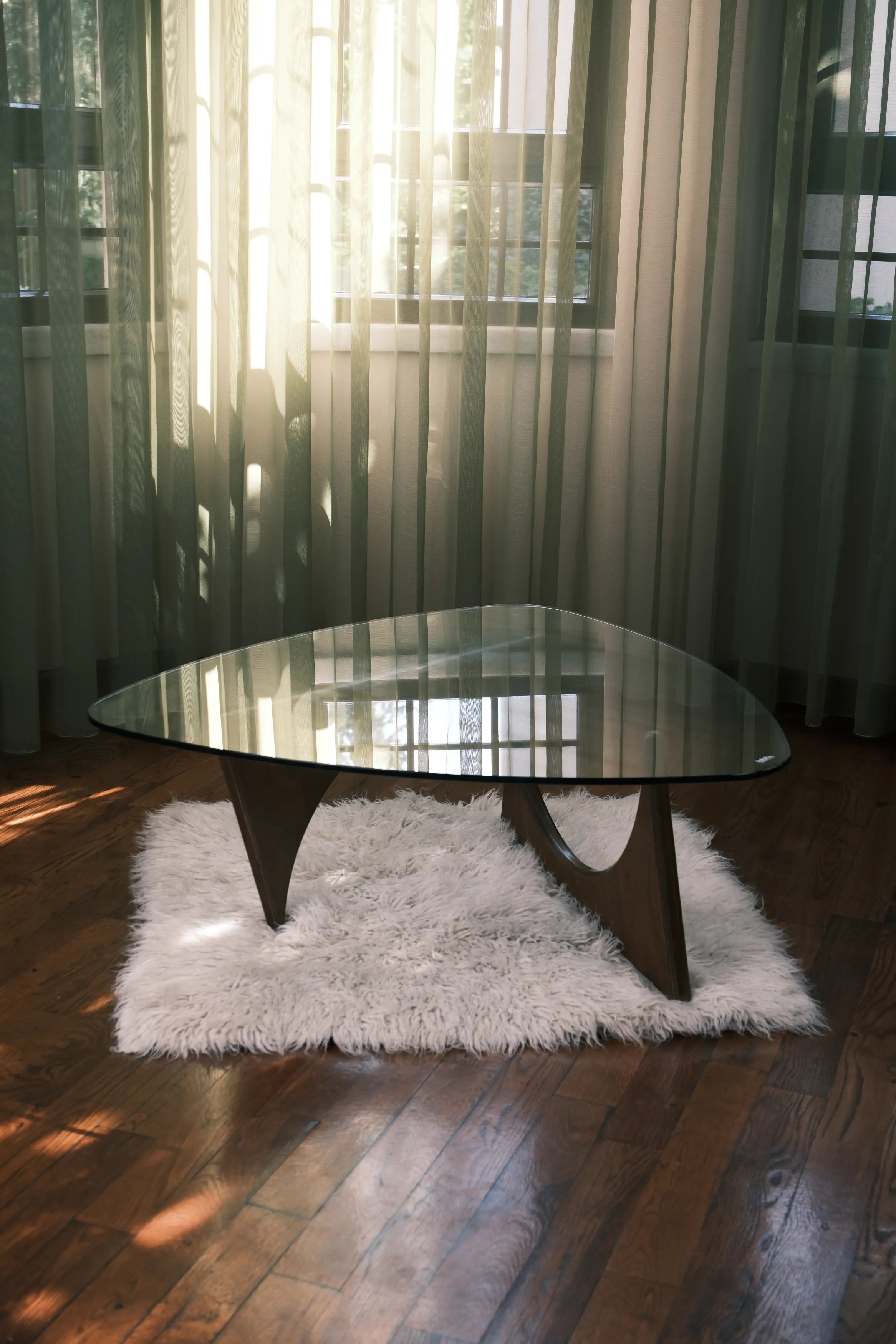 Design-Couchtisch mit Glasplatte und Holzgestell auf weißem Teppich in hellem Raum mit Parkettboden und lichtdurchfluteten Vorhängen.