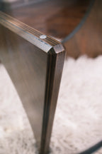 Detailaufnahme der Verbindung zwischen Glasplatte und Holzfuß eines Designer-Couchtischs auf flauschigem, weißem Teppichboden.