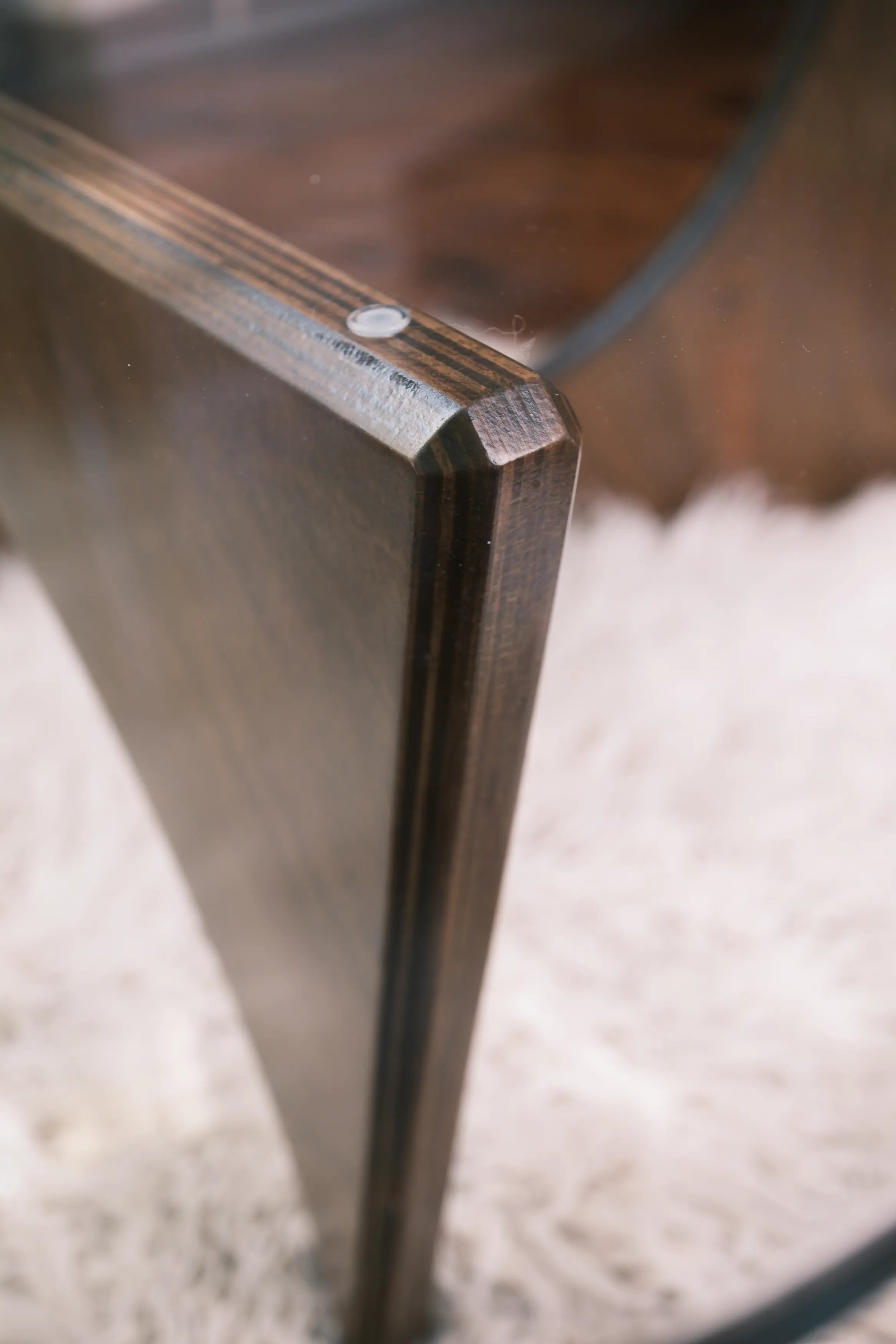 Detailaufnahme der Verbindung zwischen Glasplatte und Holzfuß eines Designer-Couchtischs auf flauschigem, weißem Teppichboden.