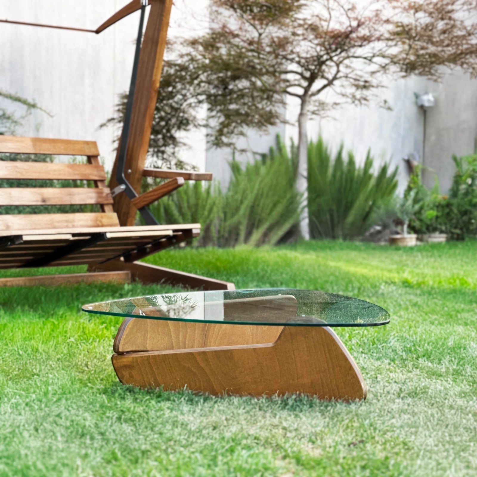 Moderner Couchtisch mit Glasplatte und geschwungenem Holzfuß im Garten neben dunklem Rattanstuhl vor weißer Hauswand