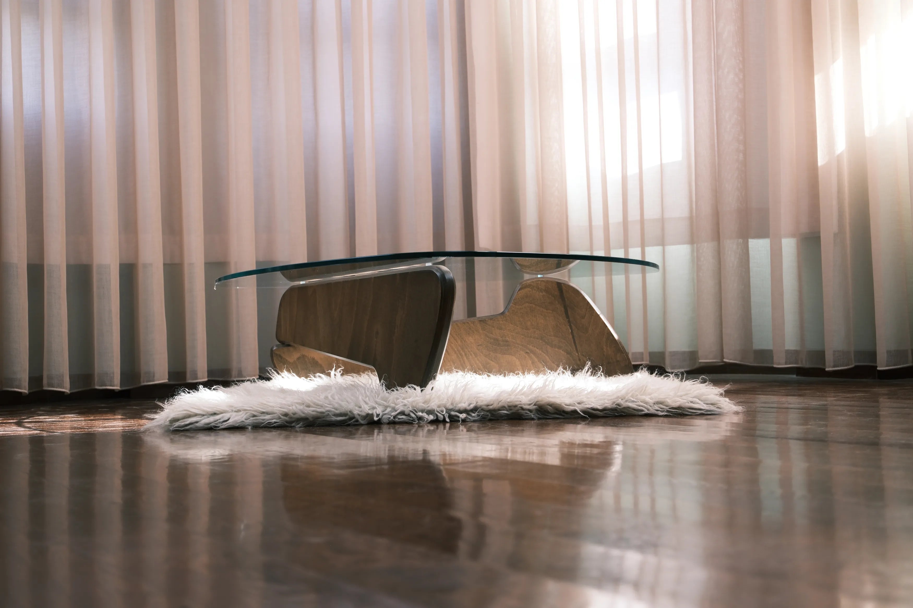 Eleganter Designer-Couchtisch mit Glasplatte und markantem Holzfuß auf flauschigem Teppich vor lichtdurchfluteten Vorhängen.