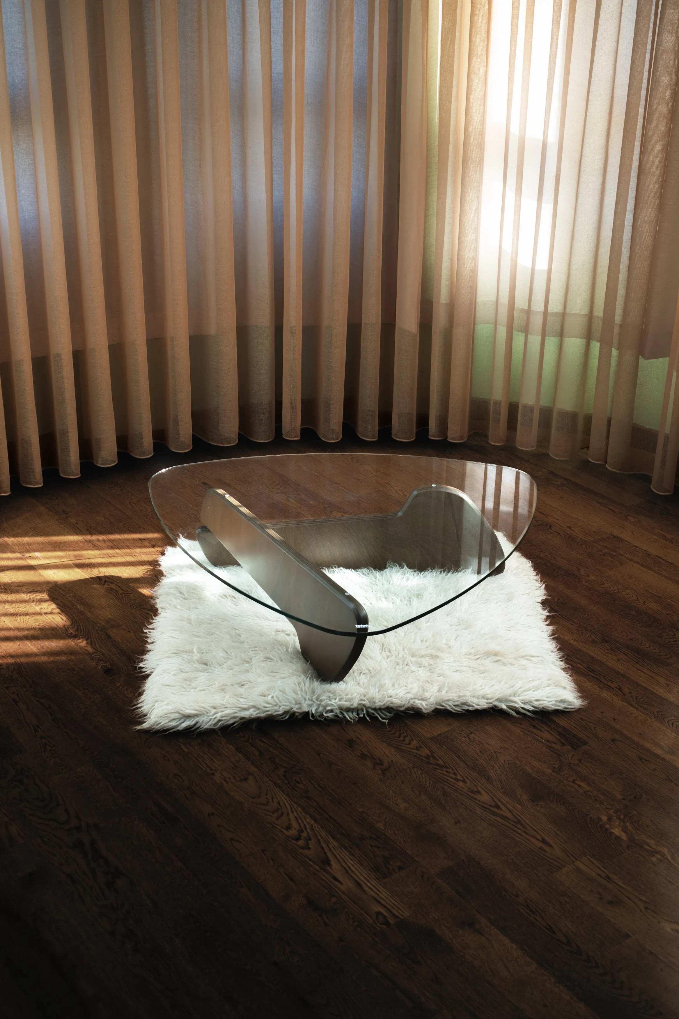 Modern geformter Glastisch mit skulpturalem Holzfuß auf weißem Teppich in sonnigem Raum mit dunklem Parkett und beigen Vorhängen.