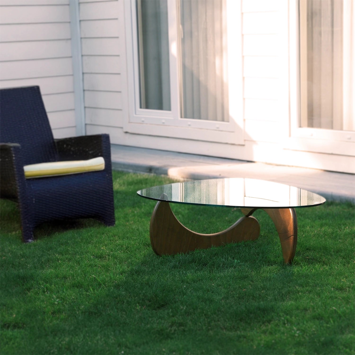 Moderner Couchtisch mit Glasplatte und geschwungenem Holzfuß im Garten neben dunklem Rattanstuhl vor weißer Hauswand