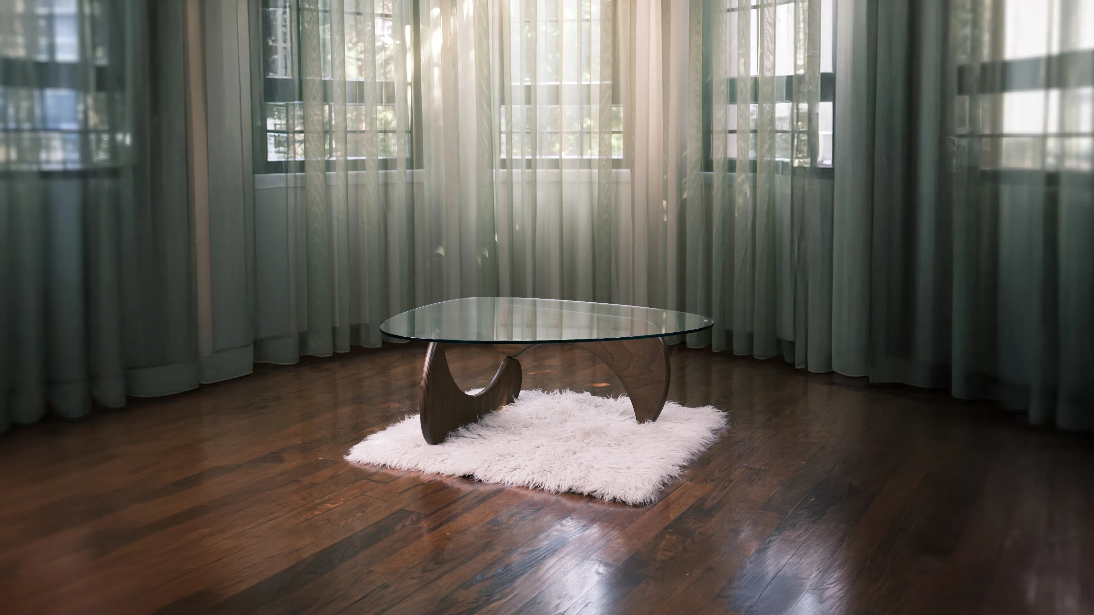 Moderner Couchtisch mit Glasplatte und geschwungenem Holzfuß auf weißem Teppich vor bodentiefen Vorhängen in hellem Raum.