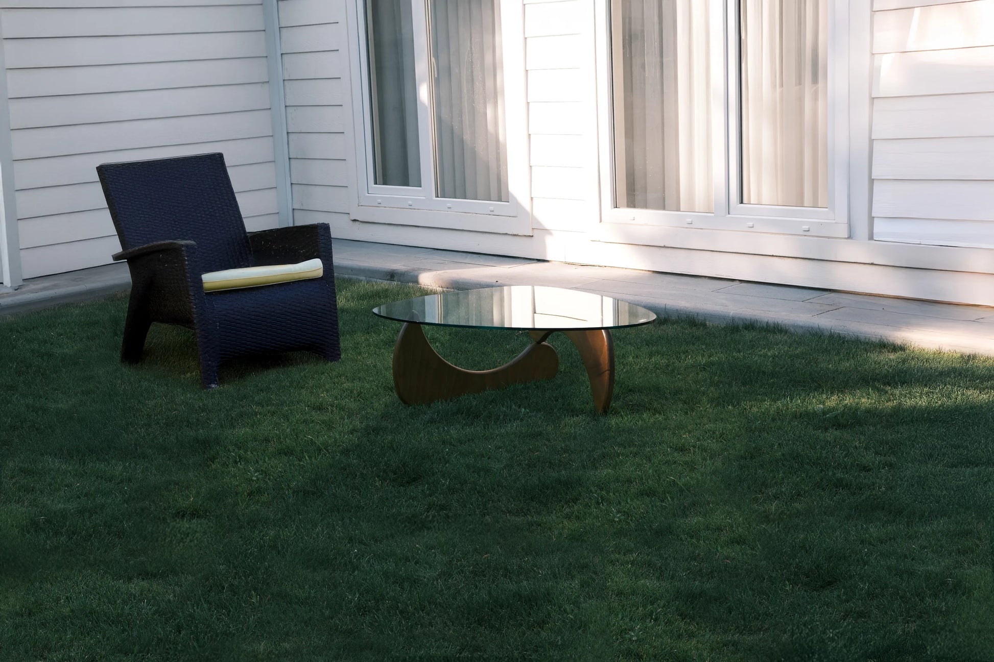 Design-Couchtisch mit Glasplatte auf Rasenfläche neben dunklem Gartensessel vor moderner Hausfassade mit großen Fenstern.